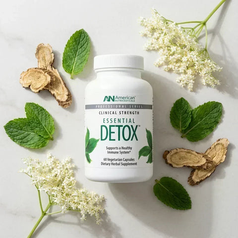 Essential Detox supplement bottle surrounded by green leaves, dried root slices, and white flowers on a pale surface