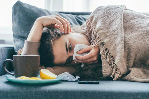 Woman lying under a blanket on a sofa, holding a tissue to her nose beside a mug and lemon wedges