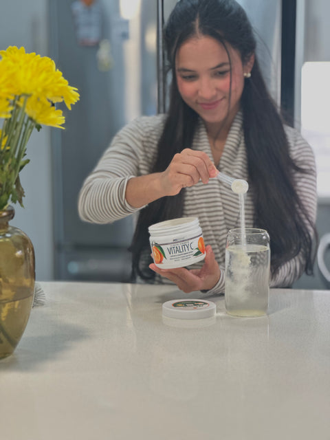 A woman in a striped sweater is shown in a kitchen setting, holding a jar labeled 'VITALITY' and using a spoon to scoop a white powder into a glass jar of water. A vase of yellow flowers is visible on the left.
