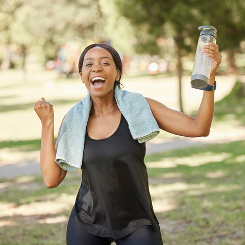A woman in a black athletic tank top and leggings celebrates in a sunny park, holding a clear water bottle and wearing a light blue towel over her shoulders.